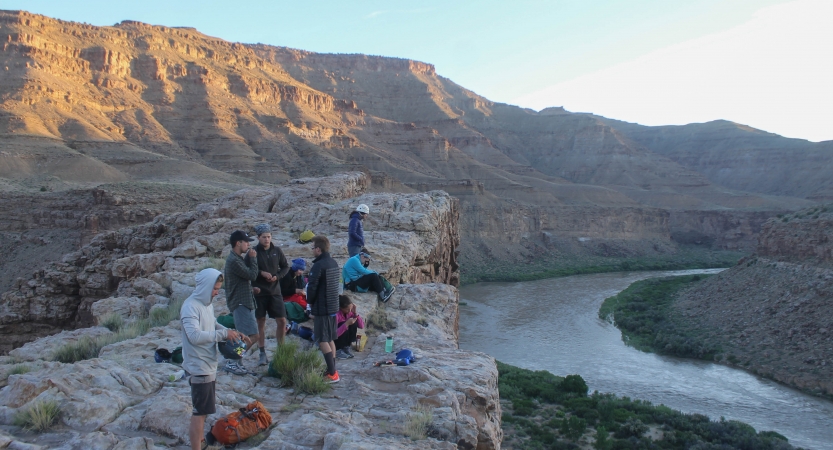 A group of people stand on a cliff, overlooking a river winding through a desert landscape.